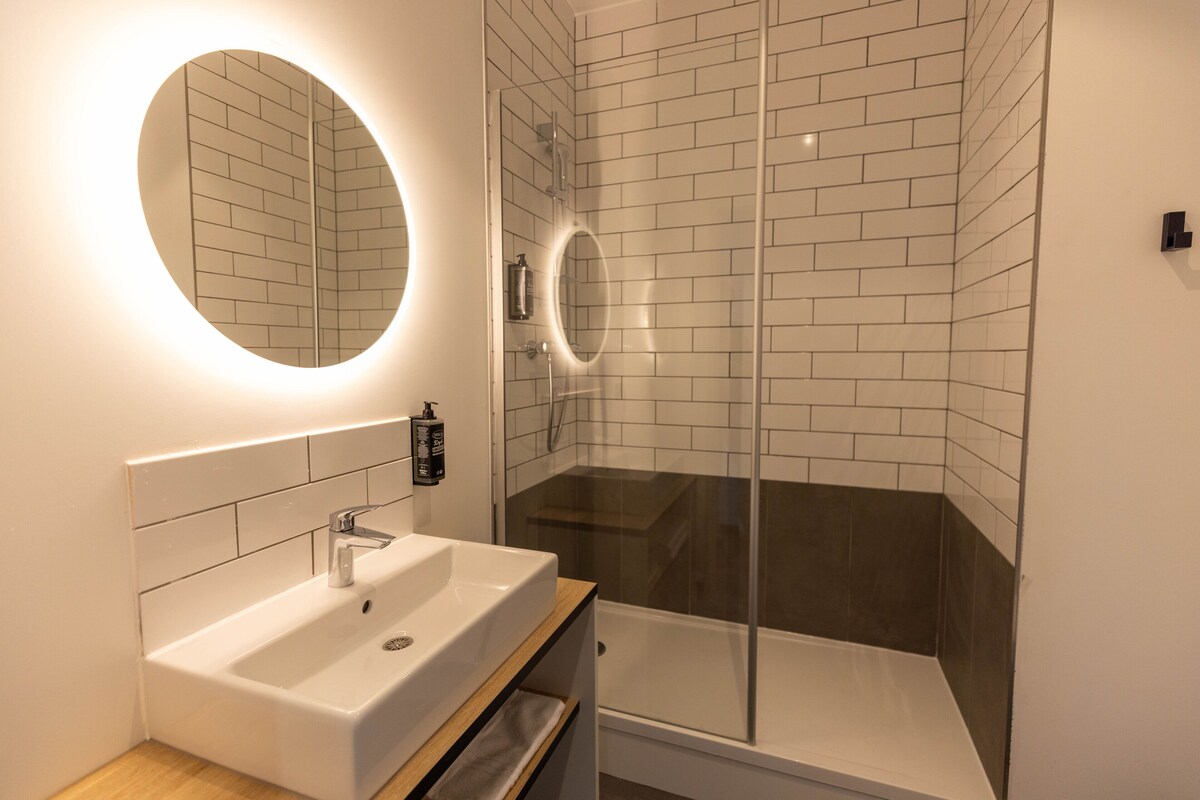 Un baño moderno con lavamanos blanco, un espejo y cabina de ducha con mampara de vidrio.