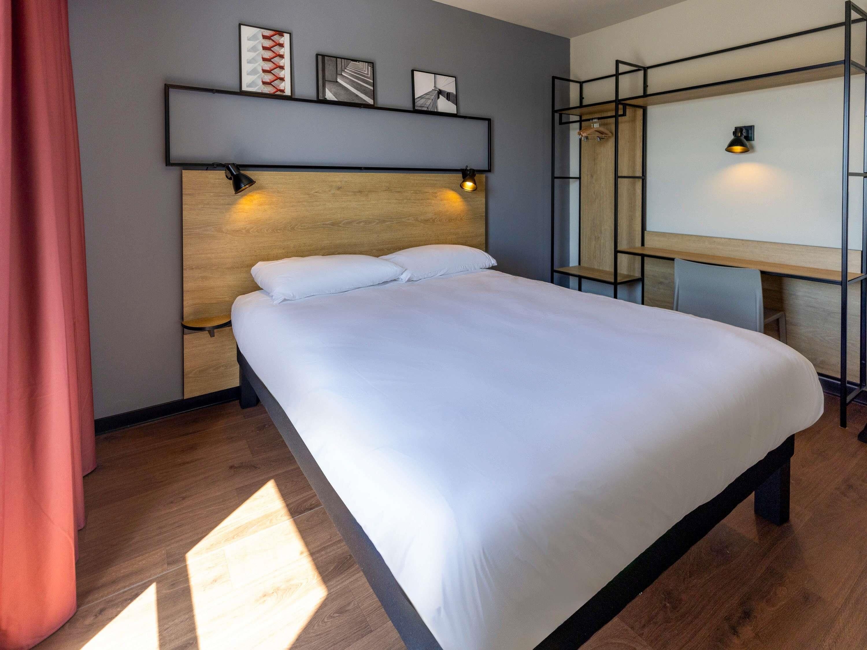 A modern hotel room with a large bed, a wooden headboard, a desk with a chair, and a wall-mounted TV.