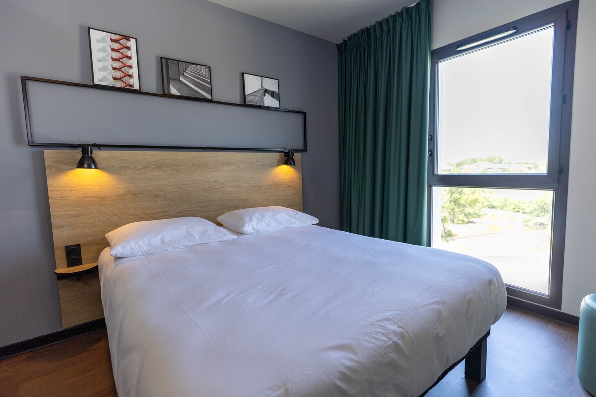 Una cama bien hecha con sábanas blancas, cabecero con dos luces fijadas a la pared, y una ventana con cortinas verdes.