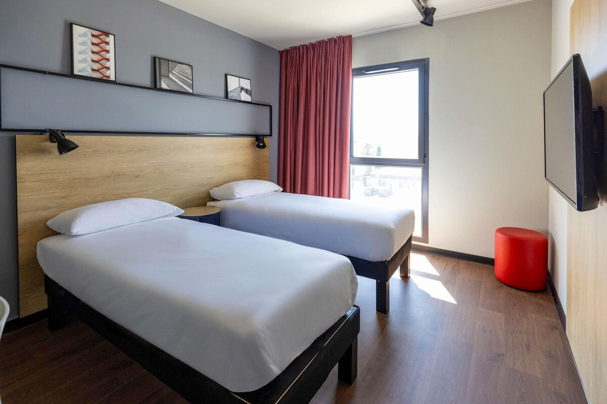Habitación de hotel con dos camas, cabecero de madera, cortina roja, taburete rojo y ventana con vistas.