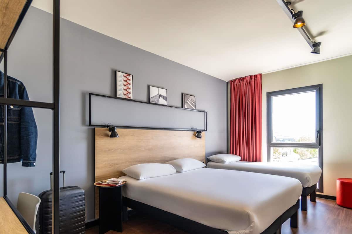 Ein modernes Hotelzimmer mit einem Holzkopfteil, zwei Betten, einem roten Vorhang und einem Fenster mit Blick.