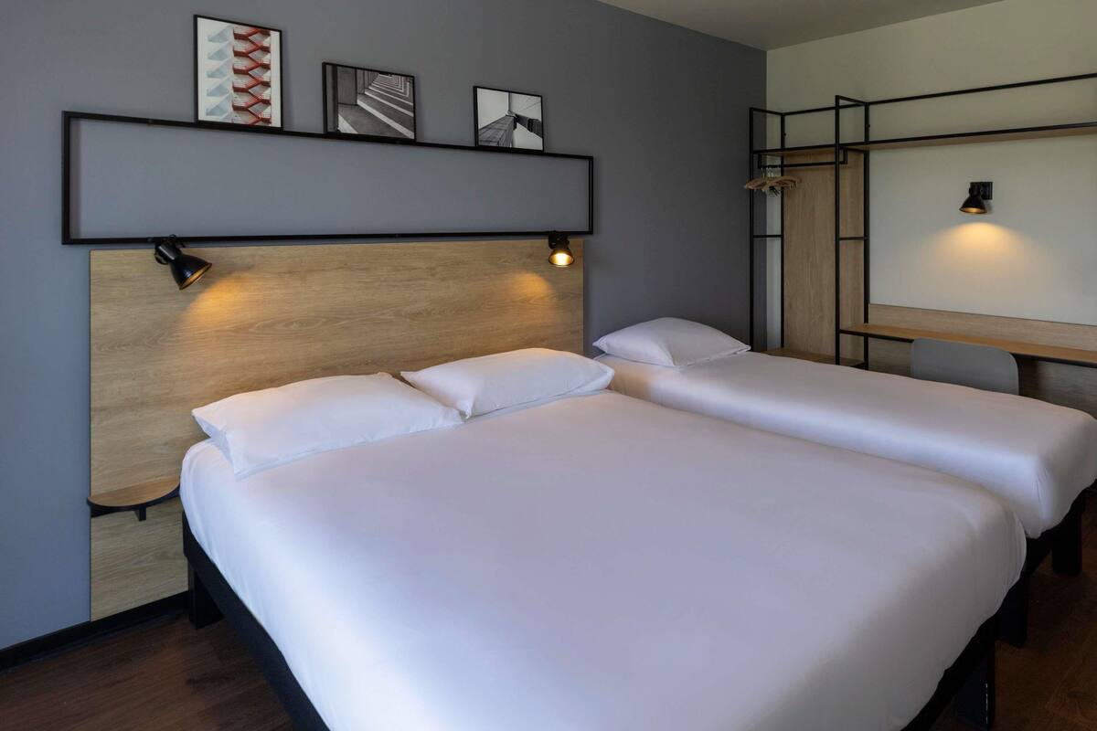 A hotel room with two beds, a wooden headboard, a shelf with framed pictures, and a desk with a lamp.