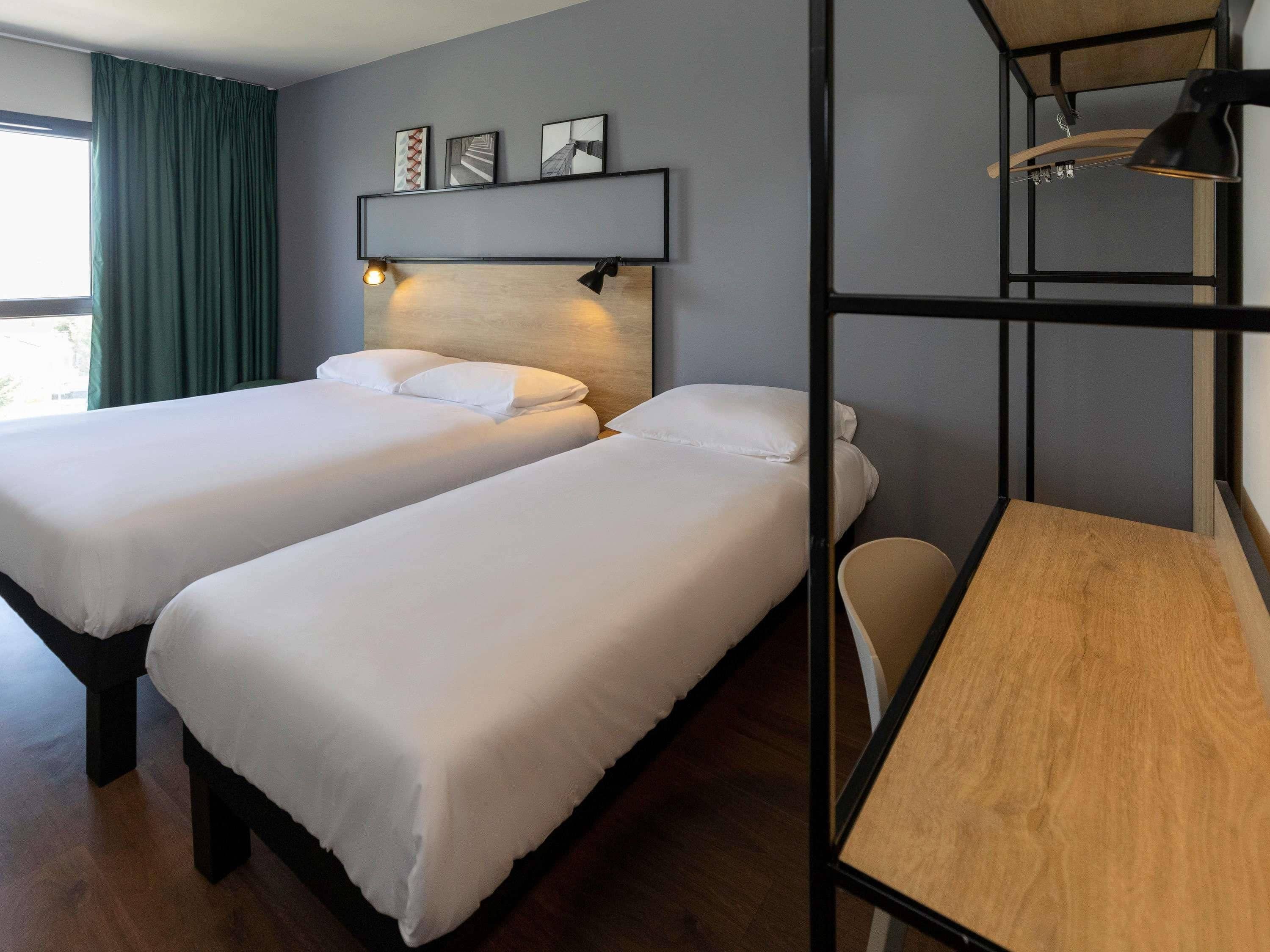 Ein Zimmer mit zwei Betten, einem Schreibtisch, einem Stuhl, einem Regal mit Bildern und einem Fenster mit Vorhängen.