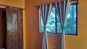 A room with a wooden door, a window with curtains, and a view of greenery outside.
