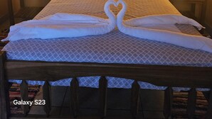 A neatly made bed with a heart-shaped towel arrangement, a painting of a tropical landscape on the wall, and two bedside lamps.