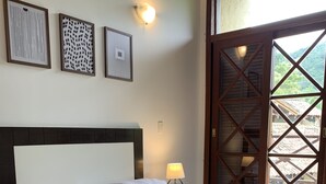 Una cama bien hecha con sábanas blancas, un edredón gris y toallas enrolladas sobre la cama. Hay una mesita de noche con una lámpara al lado. La habitación tiene un cabecero de madera y un ventanal con vistas a zonas verdes.