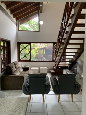 A spacious living area with a high ceiling, a wooden staircase, and large windows offering a view of the outdoors. - House H 11 in Maresias, gated community facing the beach. (Praia de Maresias São Sebastião)