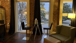 Una habitación con pared de ladrillo, un ventanal con vistas a árboles, un trípode, dos sillas y un sofá.