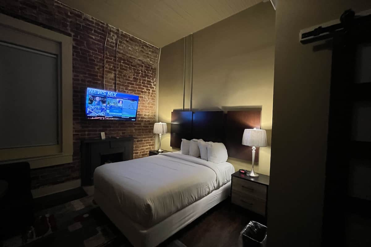 A hotel room with a brick wall, a flat-screen TV mounted on the wall, a bed with white linens, two bedside tables with lamps, and a digital clock.