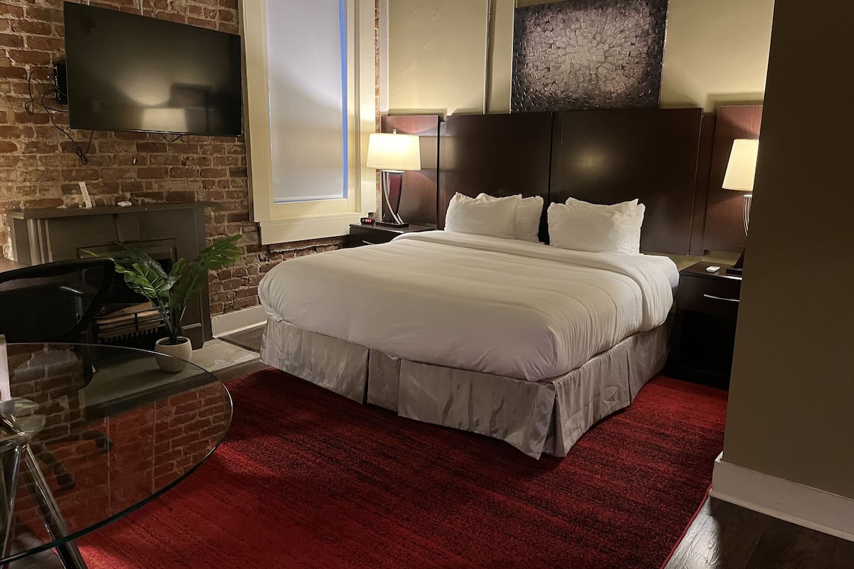 A hotel room with a large bed, a flat-screen TV mounted on a brick wall, a desk with a chair, and a red carpet.