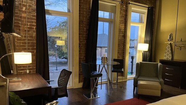 Habitación de hotel con cama, escritorio, silla, una mesita pequeña y un sofá. Amplias ventanas con cortinas, una lámpara en el escritorio y una planta en el suelo.