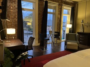 Una camera d'albergo con un letto, una scrivania, una sedia, un tavolino e un divano. Grandi finestre con tende, una lampada sulla scrivania e una pianta sul pavimento.