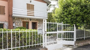 Ein Gebäude mit einem steinernen Mauerwerk, einem weißen Balkon und einer üppigen, grünen Hecke, dessen Eingang durch ein Tor gesichert ist.
