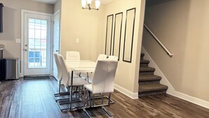 Un espace repas avec une table et des chaises blanches, un lustre, un escalier et une porte donnant sur l’extérieur.