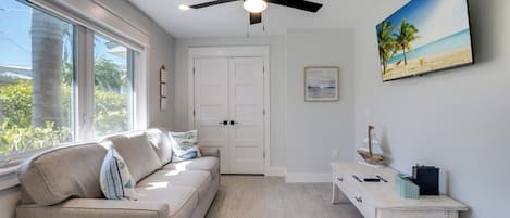 Et værelse med ventilator, sofa med puder, et lille bord med pynteting, et billede i ramme på væggen og en dør med udsigt til grøn natur udenfor.