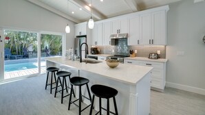 Una cocina moderna con armarios blancos, una isla grande con taburetes, horno empotrado y vista a una piscina exterior.