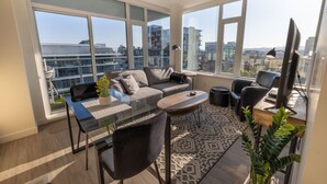 Una sala de estar moderna con una mesa de vidrio, sofá gris y vistas de la ciudad a través de amplios ventanales.