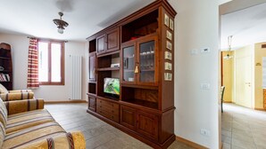 A living room with a sofa, wooden cabinet, TV, and a window with curtains. - GuestHost - Apartment located on the first floor of a building, divided into two levels without a lift.In 85 square meters of space it comfortably accommodates four people with two bedrooms located on the second floor. There is an internal staircase.Loca (Venezia)