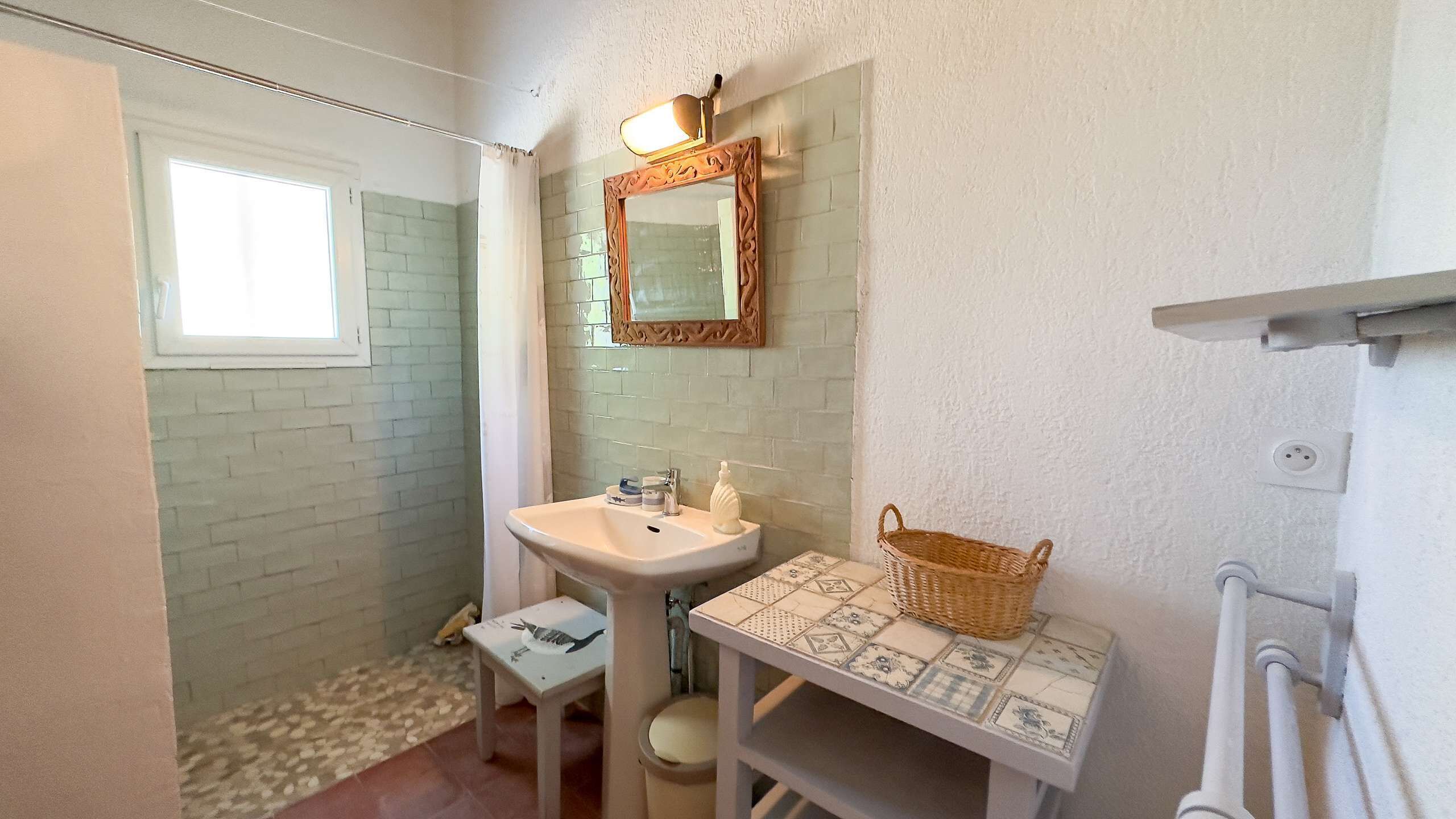 Une salle de bain avec un lavabo blanc, un miroir, une petite table avec un panier et une fenêtre.