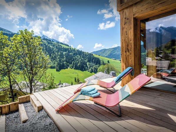 Terraza de madera con sillones de descanso y vistas a un valle verde y montañas.