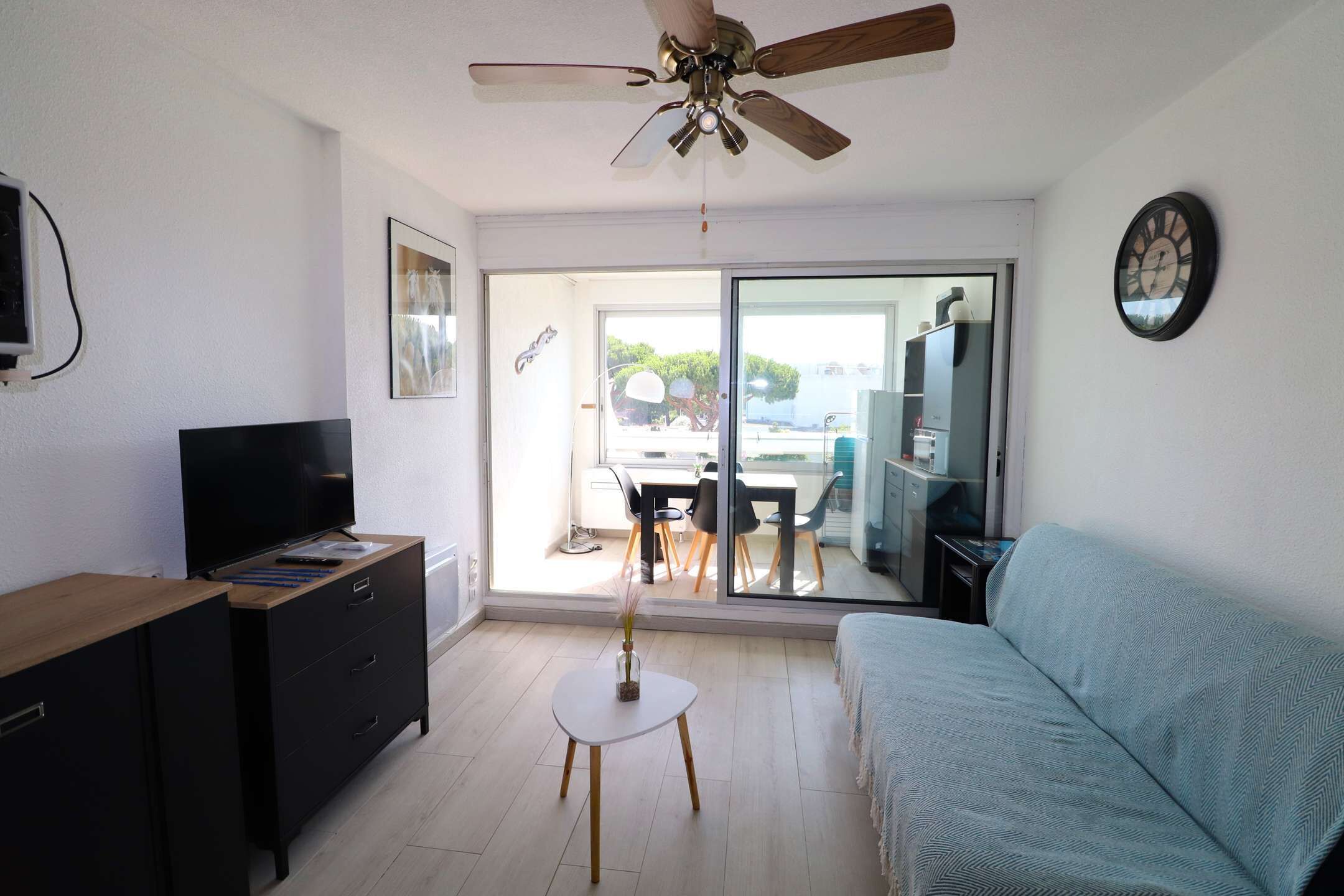 Ein modernes Wohnzimmer mit Deckenventilator, Flachbildfernseher, Sofa, kleinem runden Tisch mit Vase und einer Schiebeglastür, die auf einen Balkon führt.