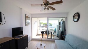 En moderne stue med ventilator, fladskærms-tv, sofa, et lille rundt bord med en vase og en skydedør, der fører ud til en balkon.