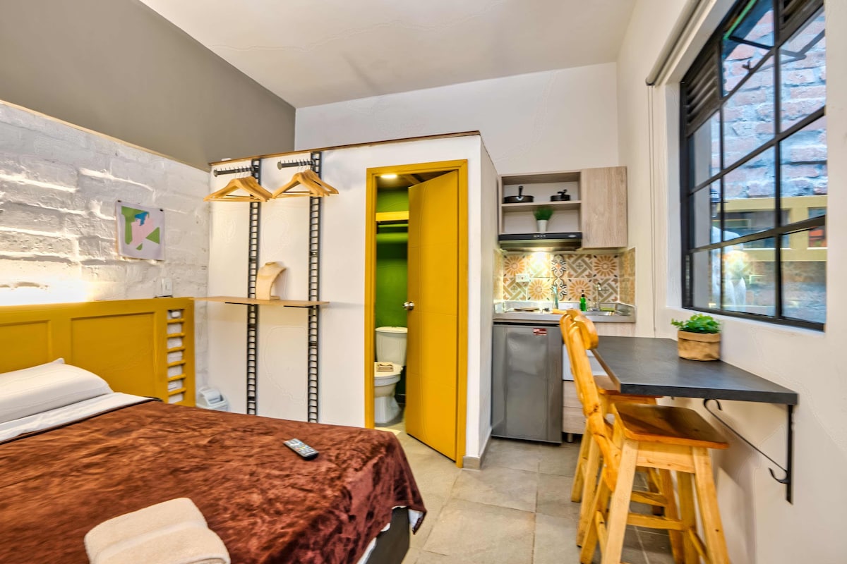 Ein kleines Hotelzimmer mit einem Bett, einer kleinen Küchenzeile und einem durchgegangene Tür erkennbaren Badezimmer.