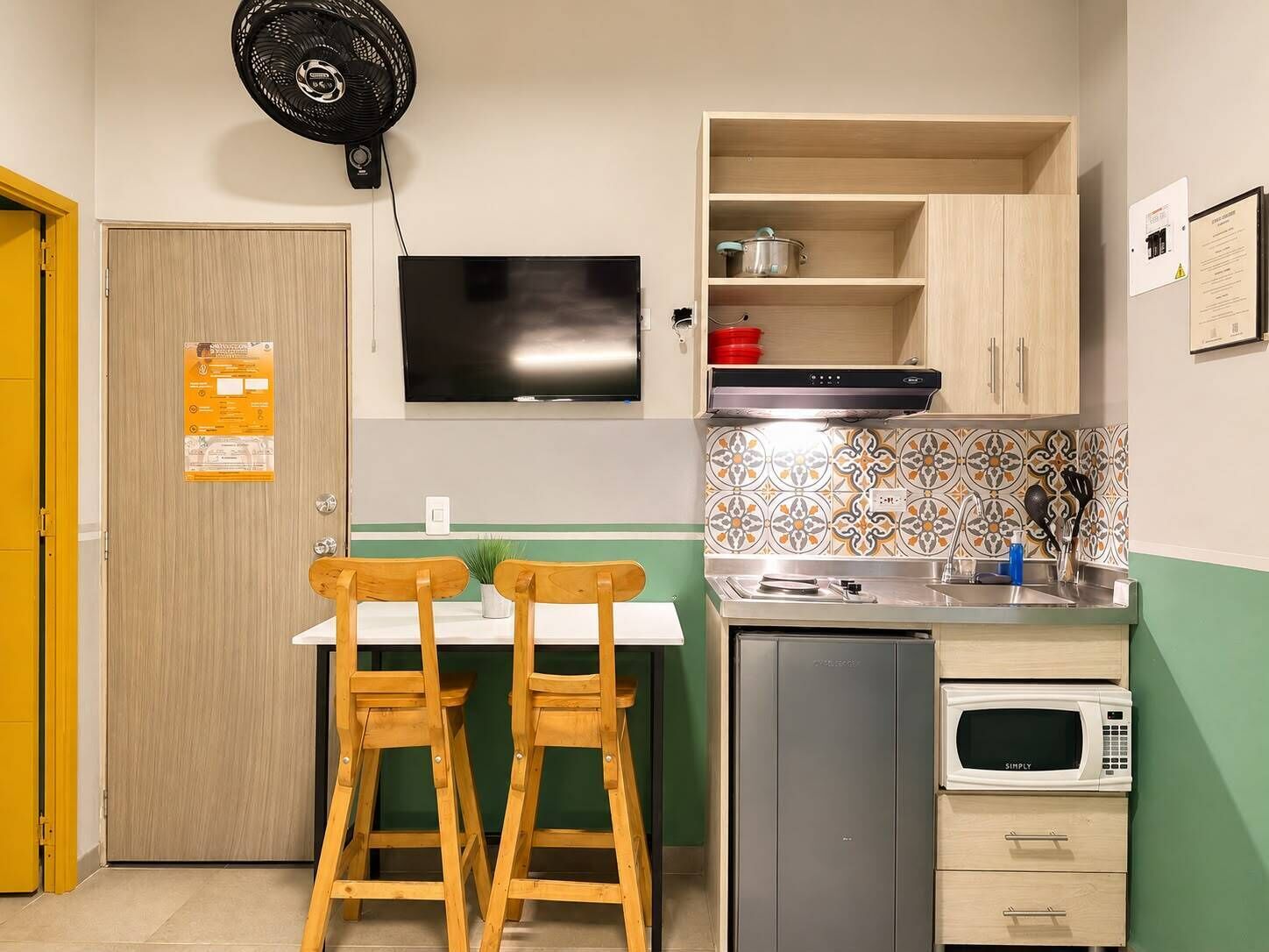 Cozinha compacta com micro-ondas, pia e um pequeno armário. Há uma TV montada na parede e um ventilador acima do balcão.
