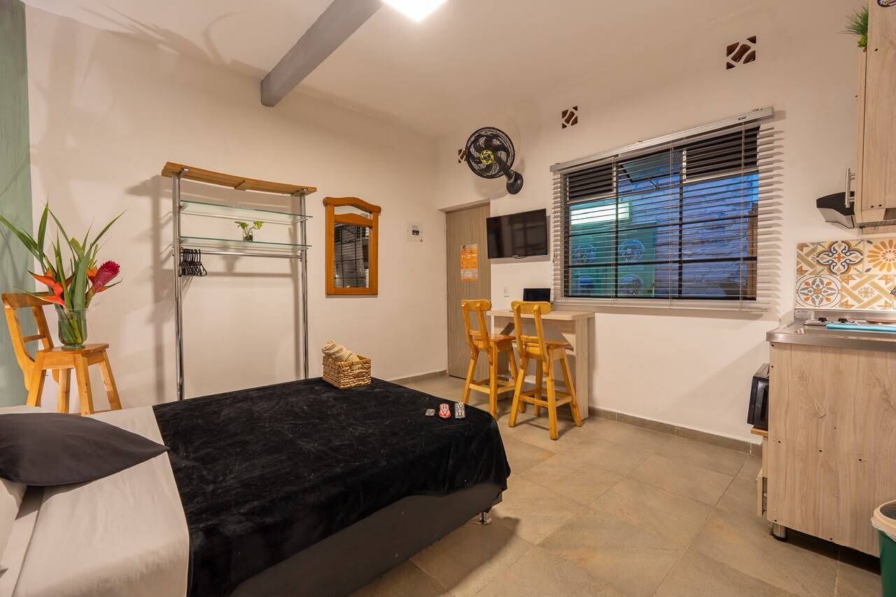 A compact room with a bed, a desk, a chair, a small table, a fan, and a window with blinds.
