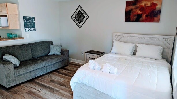 A bedroom with a bed, a gray sofa, a wooden floor, a ceiling fan, and a wall with a decorative sign. - Peaceful Studio Apt in Cental MB (Myrtle Beach)