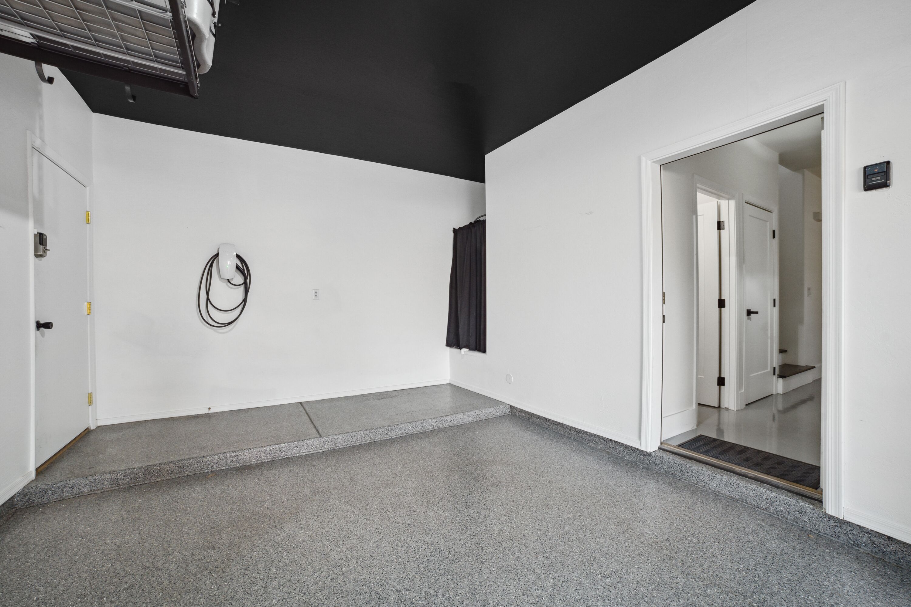 Ein Flur mit weißer Wand, schwarzer Decke und grauen Boden. Eine Tür befindet sich rechts, eine Wandleuchte links.