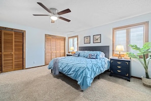 A bedroom with a bed, a nightstand, a ceiling fan, a window with blinds, and a wooden closet.