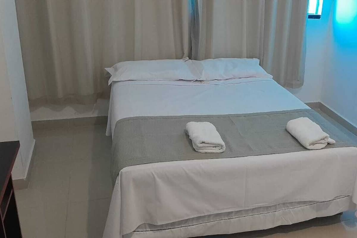 A single bed with white linens, a grey blanket, and two rolled towels.