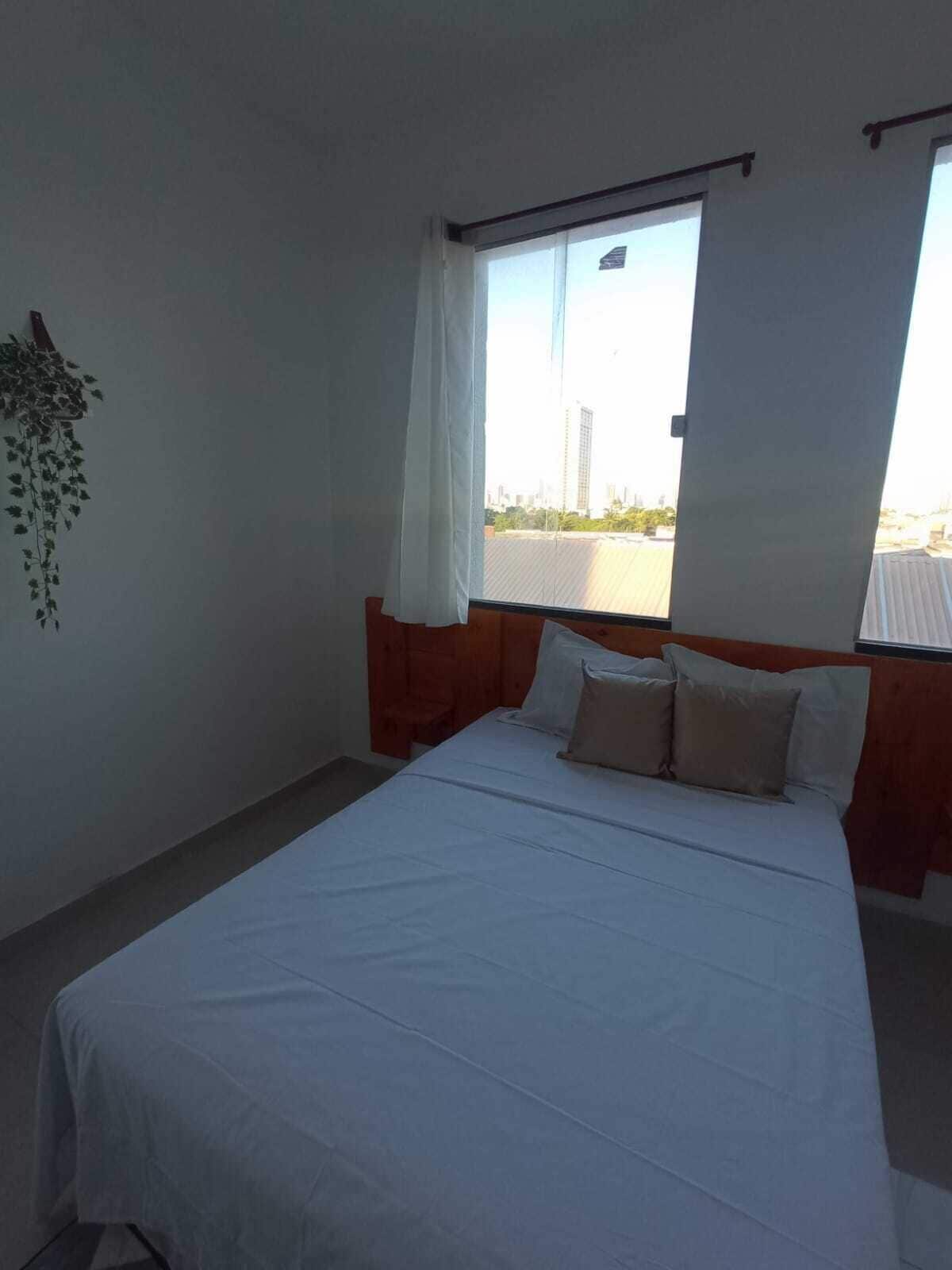 A hotel room with a bed, a window with curtains, a hanging plant, and a view of the cityscape.