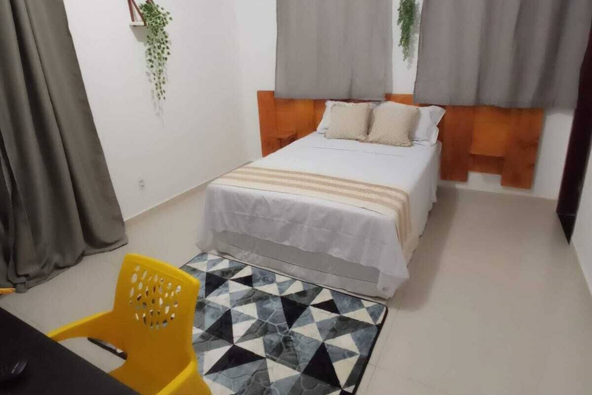 A bedroom with a bed, curtains, a small table, a chair, and a geometric rug.