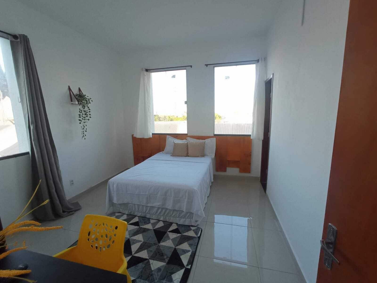 Una habitación pequeña y limpia con una cama, una silla y una mesa. Dos ventanas dejan entrar luz natural.