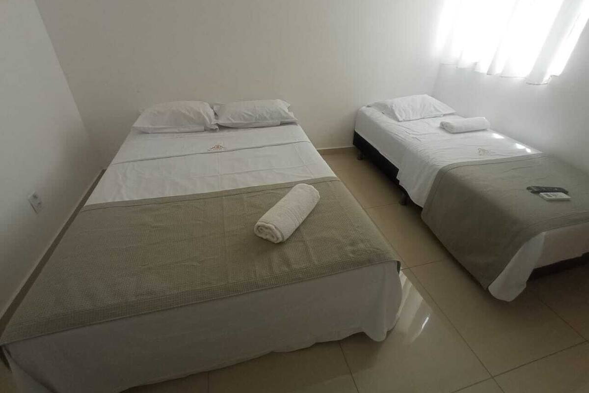 A room with two single beds, each with white bedding and pillows, a small table with a folded towel, and a window with sheer curtains.