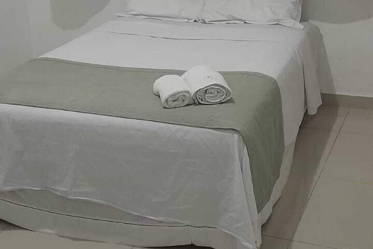 A single bed with a green bedspread and white linens, a small table with a rolled towel, and light-colored curtains.