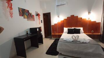 Camera da letto moderna con testiera in legno, letto con copriletto bianco e cigno di asciugamani, una scrivania con un computer e una piccola pianta in vaso.