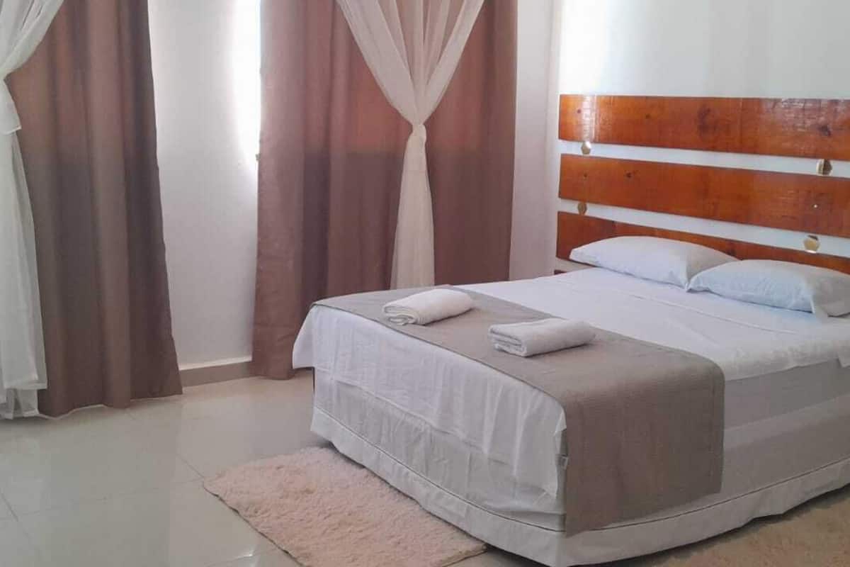 A neatly made bed with white bedding and a wooden headboard, a small bedside table, and a pink rug on a tiled floor.