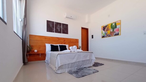 Un dormitorio moderno con cabecera de madera, cama con ropa blanca, mesita de noche pequeña, alfombra gris y un cuadro abstracto colorido en la pared.