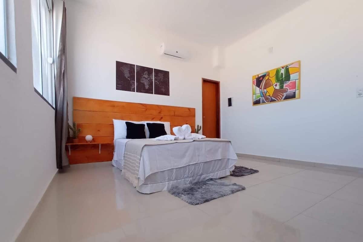 A modern bedroom with a wooden headboard, a bed with white linens, a small nightstand, a gray rug, and a colorful abstract painting on the wall.