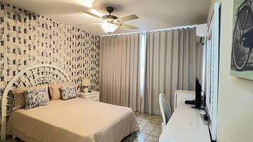 A bedroom with a bed, a desk, a chair, a ceiling fan, and a window with curtains.