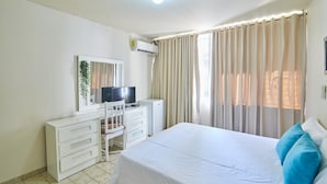 A bedroom with a bed, a desk with a TV, a dresser, and a window with curtains.