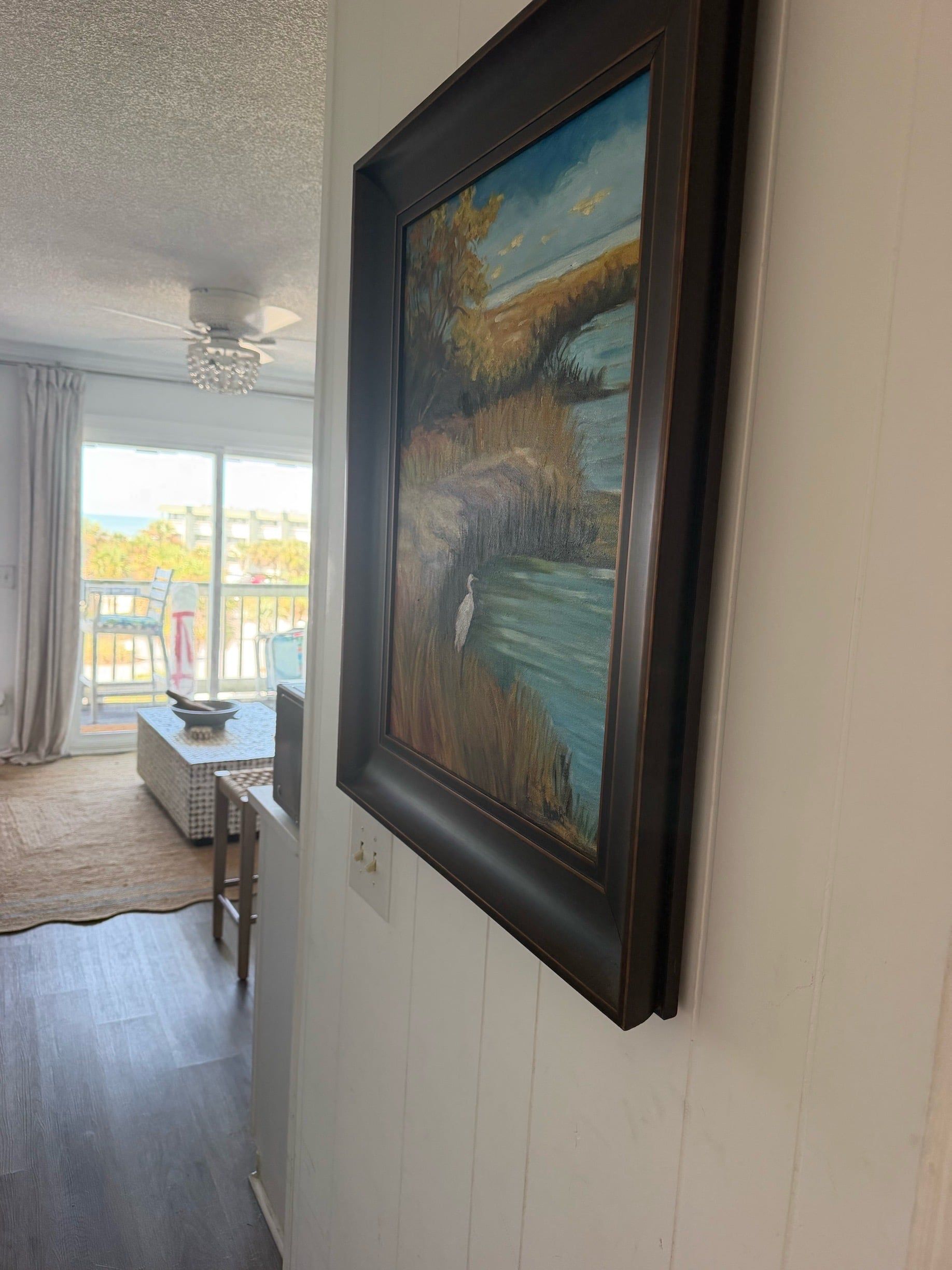 A room with a large painting on the wall, a ceiling fan, and a view through a window.