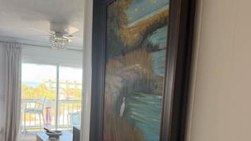 A room with a large painting on the wall, a ceiling fan, and a view through a window.