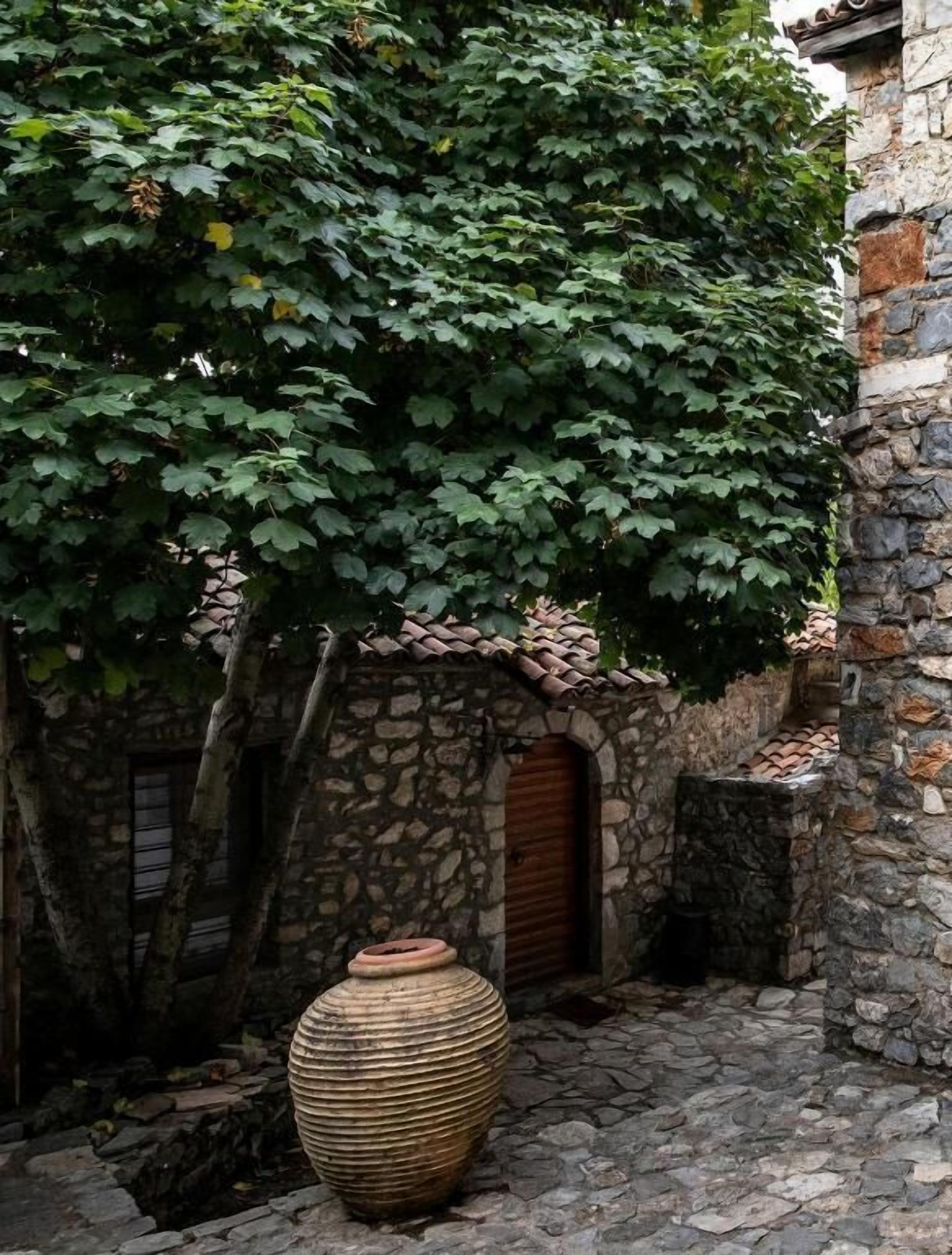Un edificio de piedra con techo de tejas, un jarrón grande y un árbol de hojas verdes.