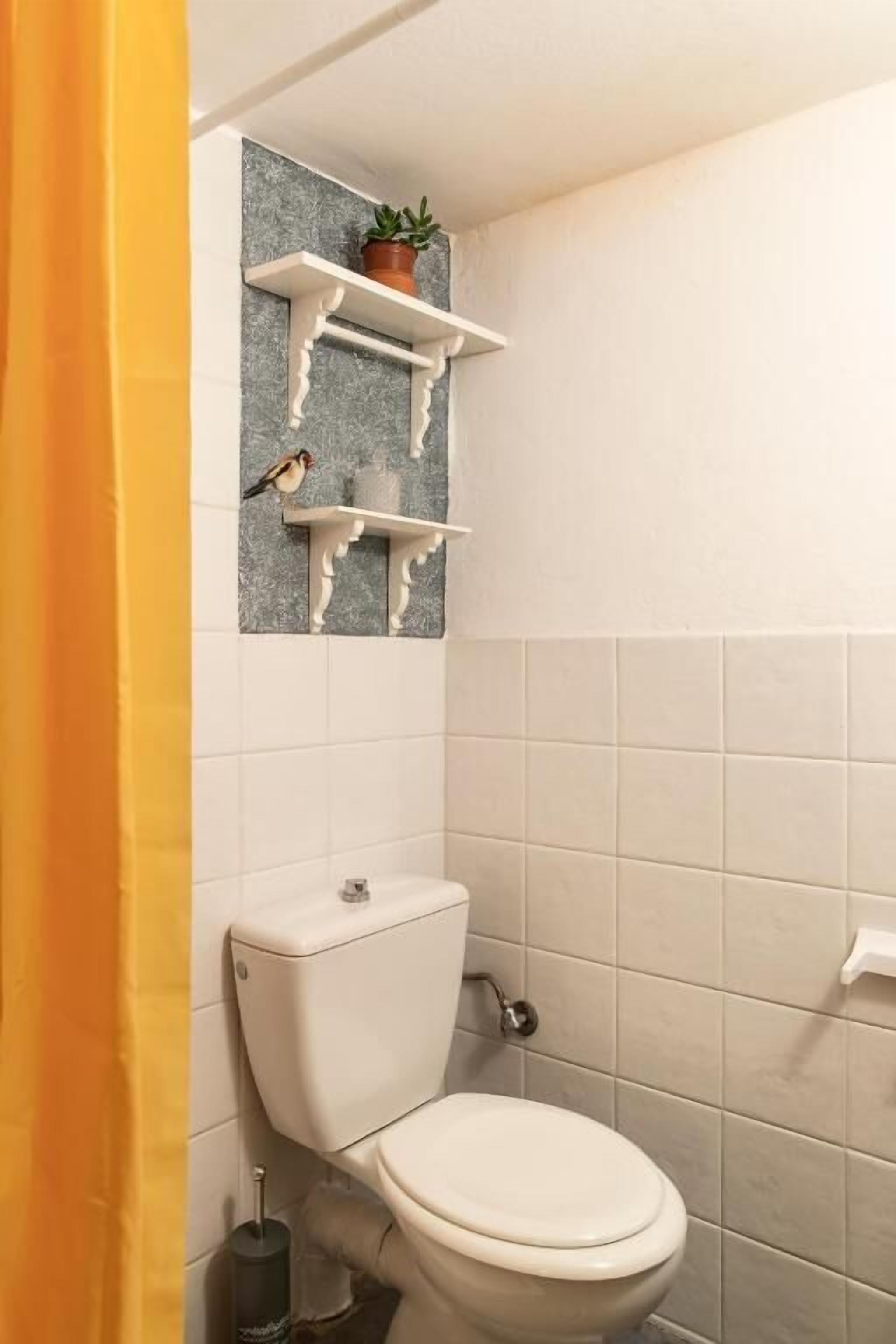 Une salle de bain avec des toilettes, des murs recouverts de carreaux blancs et une étagère sur laquelle se trouvent un pot de fleurs et une statuette d'oiseau.