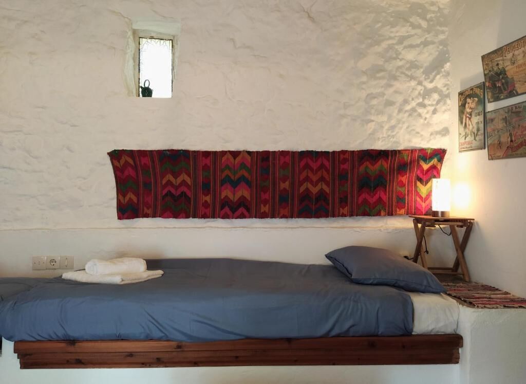 A single bed with a blue mattress and pillows, a small window, and a wall-mounted shelf with a lamp.