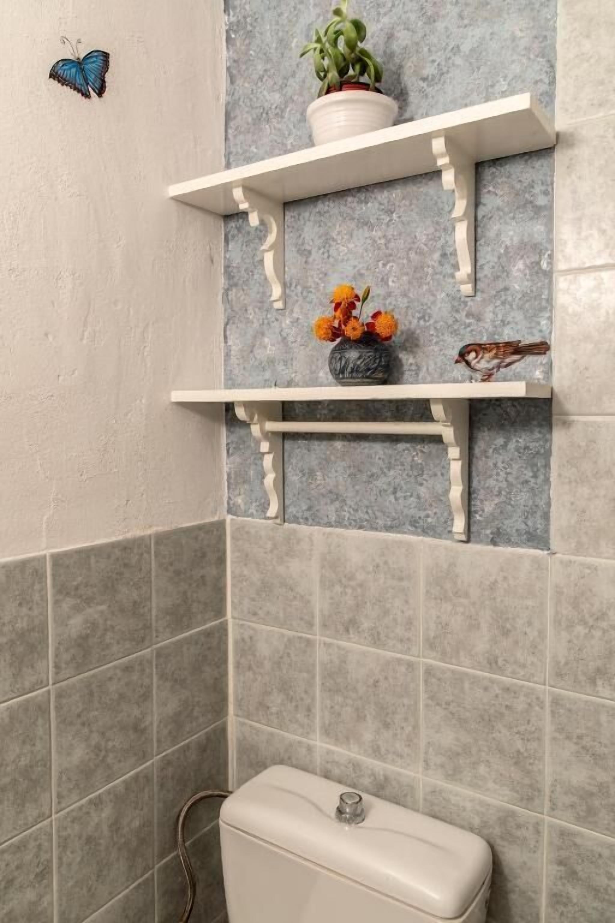 Une salle de bain avec des toilettes, des carreaux gris, une étagère blanche ornée d’une plante et de fleurs, et un autocollant représentant un papillon.